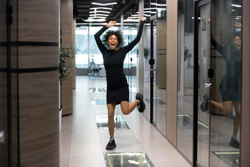 Excited mixed-race businesswoman jumps dance in office hallway raise hands scream with joy...