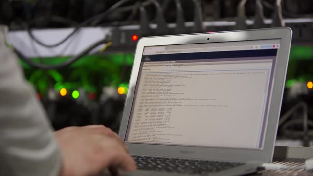 Data center diagnostics technician works and types on laptop. Mining equipment service in server room. Administrator repairs computing cluster with network hardware checking log.