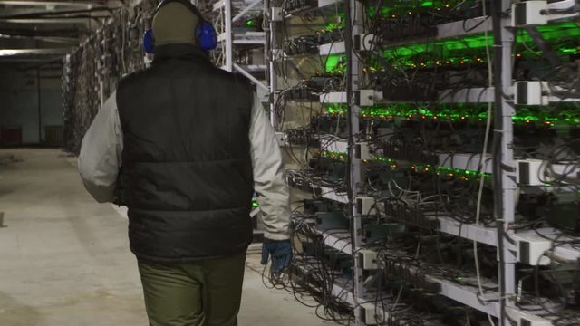 Data center diagnostics technician walks along the mining equipment racks in server room. Network administrator checks large computing cluster with network hardware. ASIC miners service engineer.