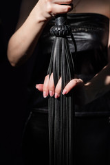 BDSM concept. A woman in a leather dress holds a black whip with a long fringe. Role-playing games for adults. Close-up of sex toys for domination.