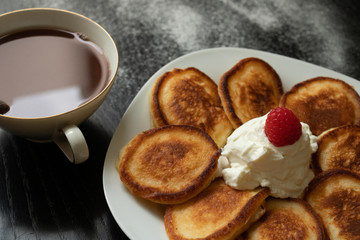 Pancakes on a white plate with raspberries and yogurt on a black background. Cup of coffee