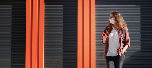 Fototapeta premium Attractive serious young woman in a mask and eyeglasses looks at the screen on a smartphone, looking for information. Against the backdrop of a closed cafe and shop during the coronavirus pandemic.