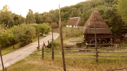 Сurved road with a small bridge. Several village buildings in the forest. Low wooden fence.