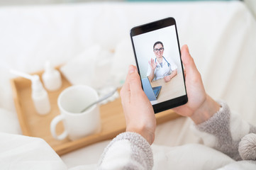 The concept of distance treatment. Rear view of a woman in bed in pajamas with a smartphone in her hands. A flu patient is watching a medical video blog.