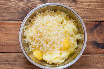 Step-by-step preparation of potato casseroles with minced meat, step 2 - grated potatoes, with eggs and cheese in a metal bowl, top view, selective focus