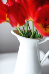 A beautiful bouquet of red tulips is standing in a white jug on the table. Festive bouquet as a gift. Focus on green stems and a jug. The concept of surprise.