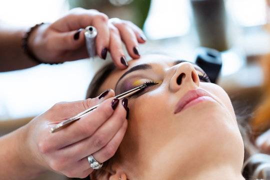 Make-up artist working with tweezers putting false long black eyelashes on female client in beauty studio.