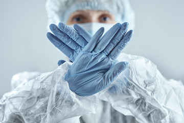 Medical worker in blur in medical mask, rubber gloves and suit makes stop sign. Confronting...
