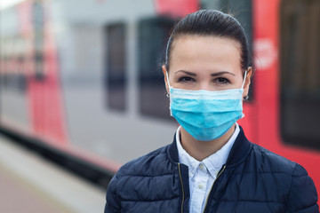 Young woman in medical protective mask on her face against coronavirus, beautiful girl standing at station near train waiting for public transport, looking at camera. Virus, pandemic Covid-19 concept