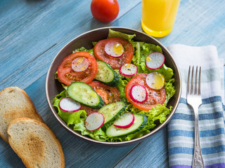 Healthy salad with fresh cucumbers, radishes, tomatoes and lettuce, olive oil and ground pepper in a plate on a wooden blue background