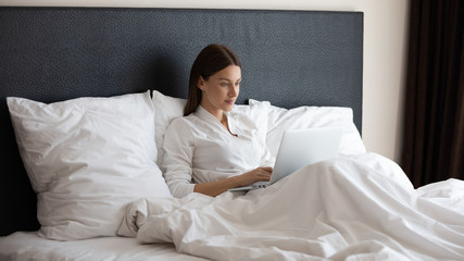 Millennial 30s woman settled comfortable sitting on bed leaned on pillows in early morning holds on lap laptop using internet web sites, check email, browse e-commerce, enjoy free time weekend concept