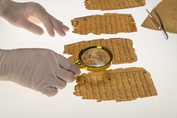 A researcher studies Arabic writing from the Koran using a magnifying glass and a table with a...