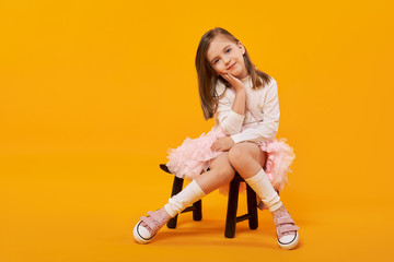 Lovely young girl sitting on small wooden chair in tulle skirt, white pullover and white gaiters