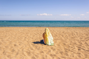 Back view of lonely Muslim woman  traveler in a colorful headscarf, scarf sitting on the sand on the beach and looking on the sea.