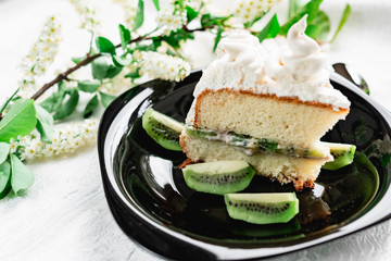 A slice of cake with meringue cream on a glossy black plate with slices of fresh kiwi. Cake decorated with a sprig of fragrant cherry blossoms