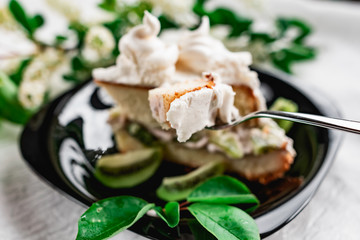 A slice of cake with meringue cream on a glossy black plate with slices of fresh kiwi. Cake decorated with a sprig of fragrant cherry blossoms