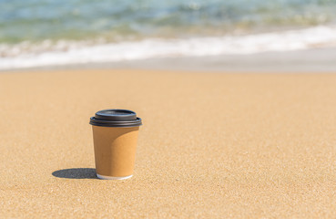 Paper mug, cup of coffee in the sand of sunny beach opposite the sea wave. summer. take away. breakfast
