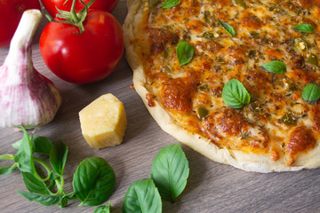 Homemade vegetarian margarita pizza with Parmesan and mozzarella cheese and traditional tomato sauce, topped with fresh green basil leaves on a wooden surface surrounded by its ingredients