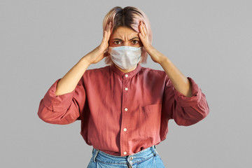 Anxious young Caucasian female holding hands on head being struck with panic wearing disposable protective face mask against airborne respiratory illness, contagious disease or industrial emissions