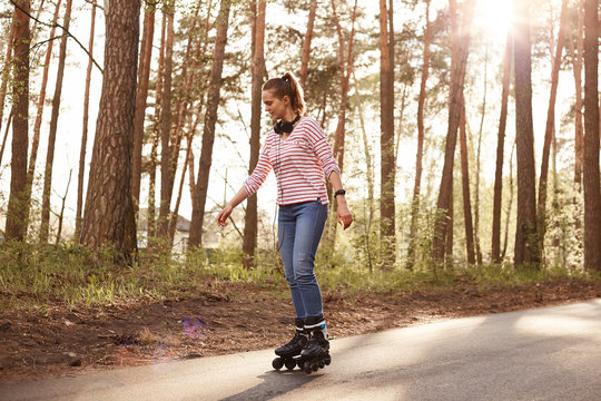 Full length photo of adorable female with ponytail and headphones around beck, woman dresses striped shirt and jeans, listening to music, enjoying songs and beautiful nature. Lifestyle concept.