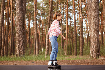 Outdoor shot of attractive woman dresses casual attire rollerblading in forest during her weekend. Lady spending time in open air, enjoying nature, being in good mood. People lifestyle concept.