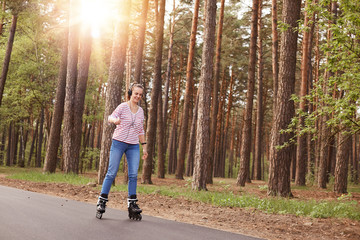 Outdoor shot of happy female spending time in open air in active way, listening to music via headphones, woman wearing casual attire rollerblading on asphalt road in forest. Lifestyle concept.