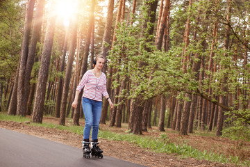 Outside portrait of active energetic young female wearing jeans and striped sweatshirt, rollerblading, heaving headphones on head, listening to music, spending free time actively, chilling out.