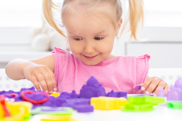 happy Girl plays kinetic sand in quarantine. Blond beautiful girl smiles and plays with purple sand on a white table.