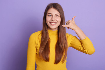 Picture of smiling lady looking directly at camera, standing against lilac studio wall and showing phone gesture with fingers. Call me. Female dresses casual yellow shirt, having long beautiful hair.