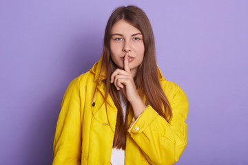 Horizontal shot of attractive woman with long beautiful hair, keeping finger near lips, calls on to keep silence, covering her mouth with fore finger,wearing casual attire against lilac wall.