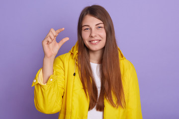 Closeup portrait of smiling dark haired woman with beautiful hair wearing yellow jacket, showing small size, looking directly at camera and smiles, posing isolated over lilac studio background.
