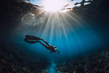 Free diver woman with fins glides over coral bottom and amazing sun rays. Freediving underwater in...