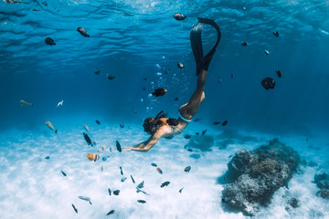 Free diver girl with fins glides over sandy bottom with tropical fishes in blue sea