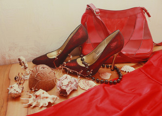Composition of shoes, a red dress, shells, a mirror, jewelry, and dried flowers on a wooden table.
