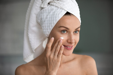 Close up face reflected in mirror of beautiful 35s woman applying cream self-care grooming activity after shower indoor. Wrinkles prevention antiaging beauty treatment for healthy radiant skin concept