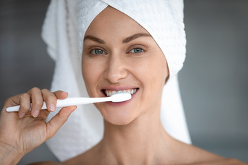 Close up face of 30s woman after shower with towel oh head holding toothbrush brushes her teeth remove plaque in morning looks at camera, caring about oral hygiene, everyday routine self-care concept