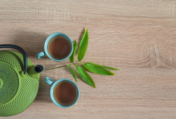 Obraz na płótnie Canvas Tea time concept in asian style. Two chinese cups of tea, iron teapot and bamboo leaves on wooden background. Space for text, top view
