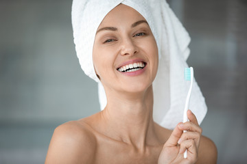 Close up fresh woman took a shower wears towel on head holds toothbrush looking at camera, has charming wide toothy smile perfect straight white teeth, oral hygiene treatment for healthy tooth concept