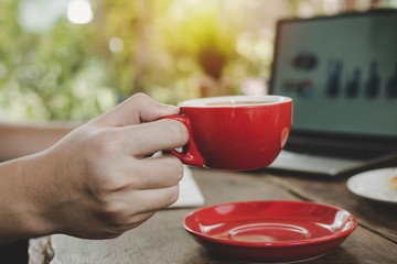 young man hand holding red coffee cup relaxed and working with digital laptop computer on desk at cafe, self quarantine, work from home, digital marketing, business finance, network technology concept