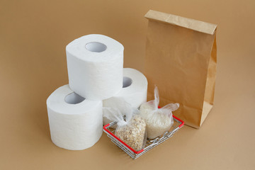 Paper bag, toilet paper and various groats in bags in a grocery basket on a brown background