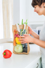 Woman holding reusable eco sackcloth fabric bag packaging with fresh organic vegetables on white kitchen table. Home delivery concept. Local farmer healthy food. Zero waste. Vertical card. Copy space.