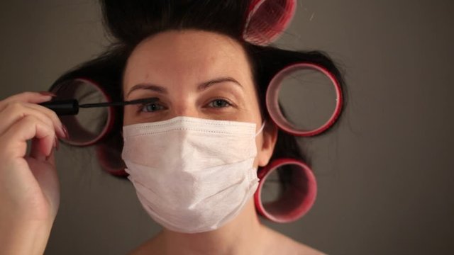 A girl with black hair in curlers and a medical protective mask paints her eyelashes and makes up. I look at the camera. Women against cold flu. Stay at home in quarantine. Macro mode