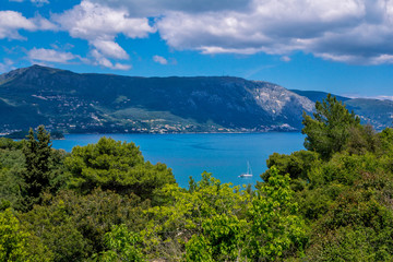 Fototapeta na wymiar Beautiful summer landscape – calm sea water surface, white yacht, green trees and mountains on the horizon. Corfu Island, Greece.