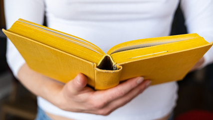 Open thick, old yellow book is held by female hands, on a blurred background.