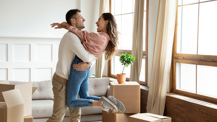 Happy young couple celebrating moving day, hugging in new apartment, loving husband lifting smiling...