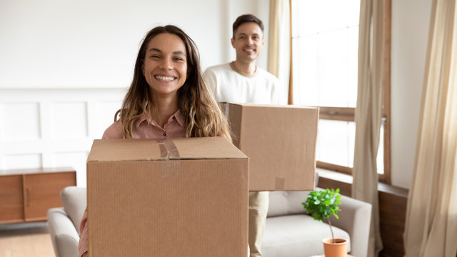 Portrait happy young couple holding cardboard boxes with belongings, moving into new first house, smiling wife and husband looking at camera, standing in living room, mortgage and relocation