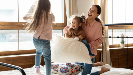 Obraz premium Happy adorable little daughters with young mother pillow fighting in bedroom, laughing and jumping, two cute preschool girls having fun with mum, playing active funny game at home