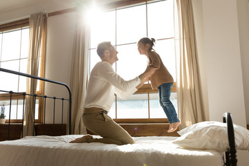 Happy father playing with little daughter, jumping on cozy bed in bedroom, smiling dad having fun with cute preschool girl, family enjoying weekend at home, spending leisure time together