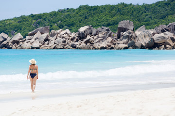 Tropical beach, turquoise blue water surrounded with huge rocks, and a walking woman in sun hat.