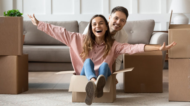 Happy Young Couple Unpacking Belongings, Relocating Into New House, Having Fun On Moving Day, Smiling Wife Sitting In Cardboard Box And Laughing In Living Room, Funny Family Activity At Home